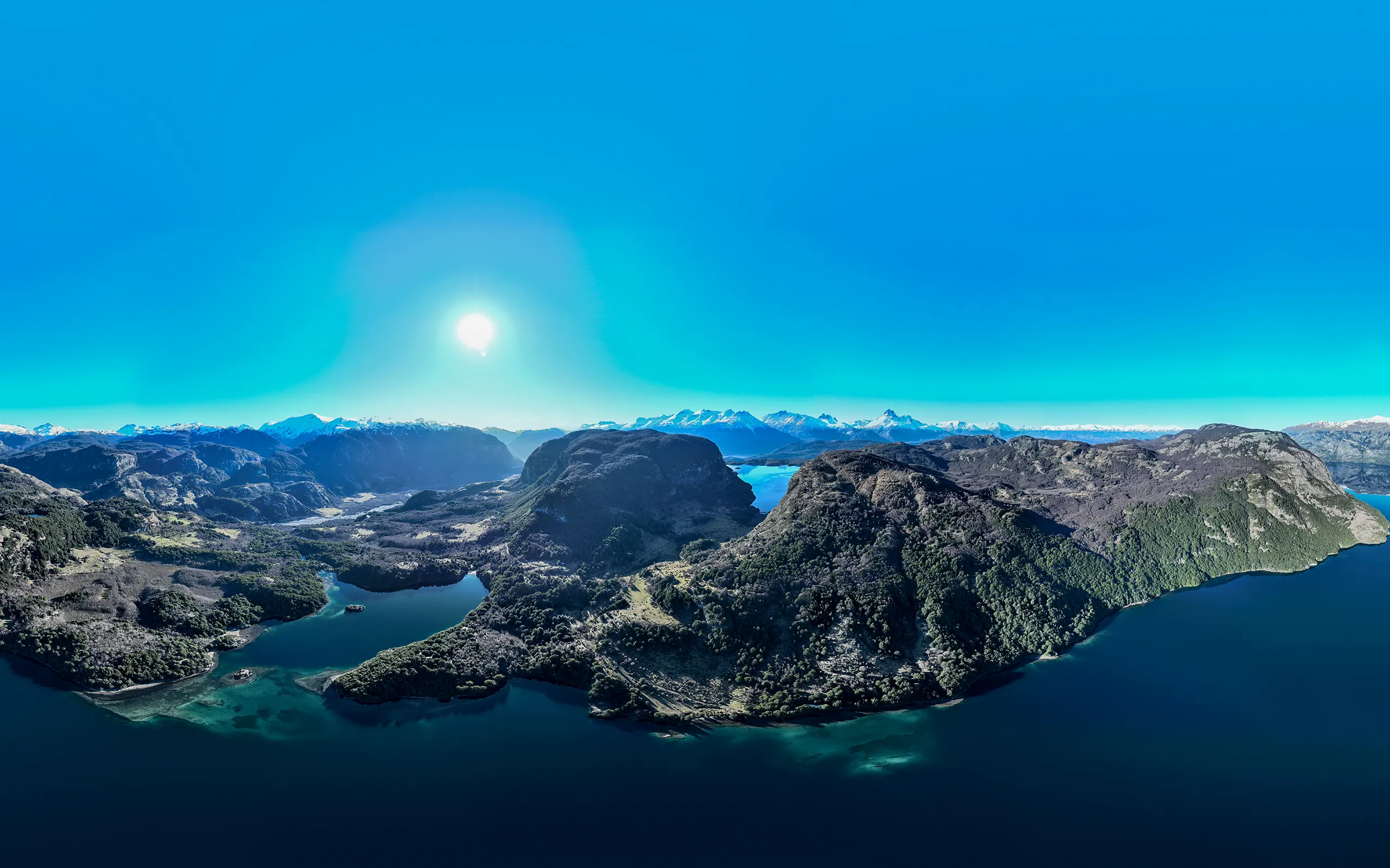 Panorámica aérea de la Patagonia chilena: una península de bosque nativo entre aguas azules, con bahías y ensenadas recortando la orilla y la cordillera nevada en el horizonte